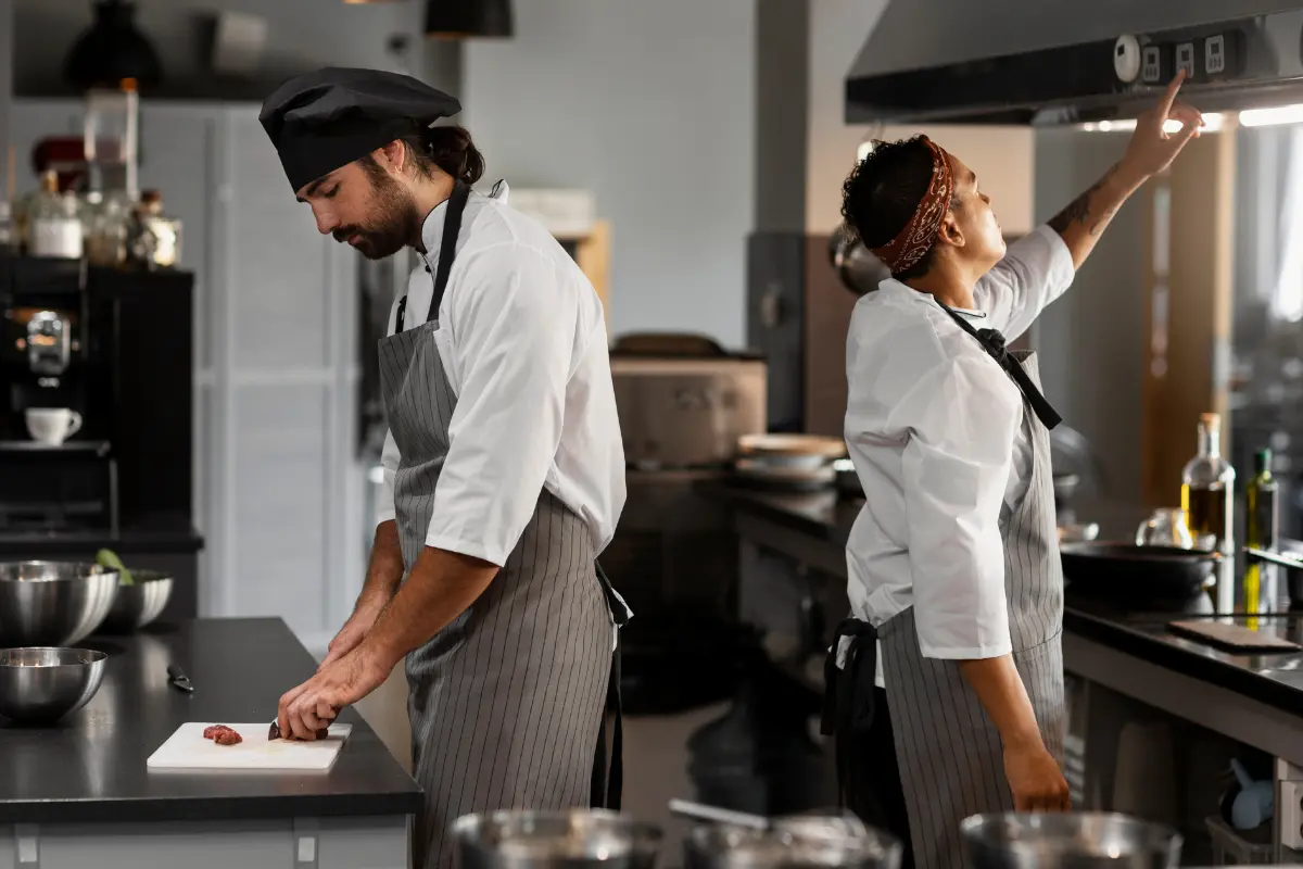 This photograph shows two professional chefs working side-by-side in a commercial kitchen, engaged in different tasks.