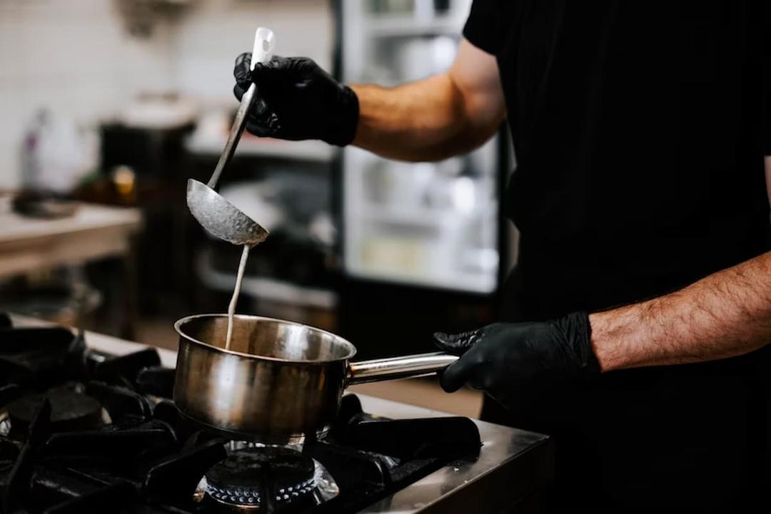 The image shows a person cooking in a professional kitchen. They are holding a stainless steel pot over a gas stove with one hand, while using a ladle to stir or pour a liquid mixture, possibly soup, sauce, or batter. The individual is wearing black gloves, which suggests a focus on hygiene and safety.