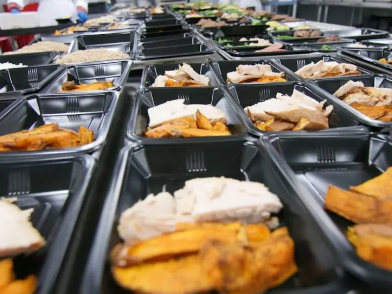 The image shows a long row of prepared meal containers arranged in a professional kitchen or large-scale food production facility. The containers are black plastic trays, neatly lined up across the entire prep table.