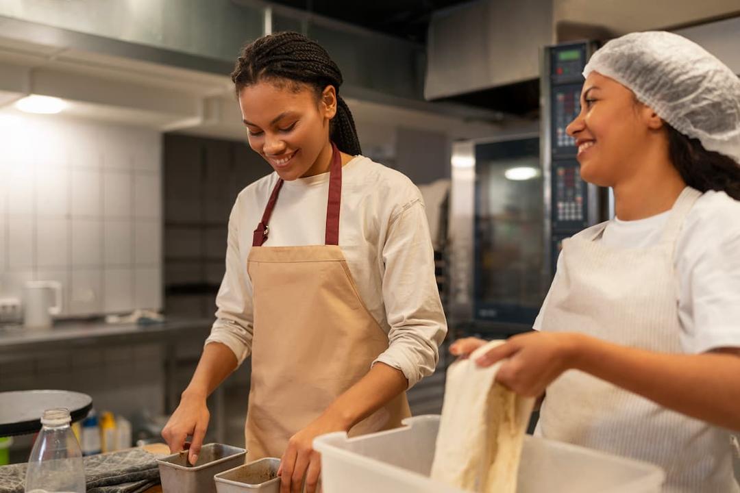 Two women smiling while baking in a commercial kitchen, illustrating how to choose the best shared use kitchen for your business.