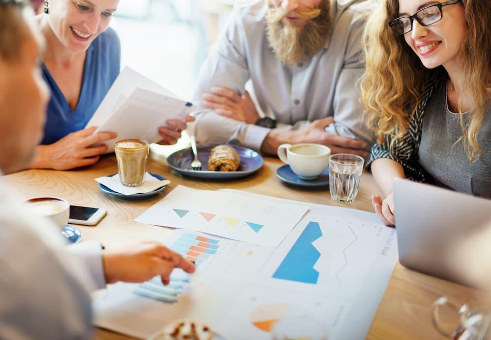 Group of professionals discussing reports and charts in a meeting, with coffee and food on the table, in a collaborative work environment